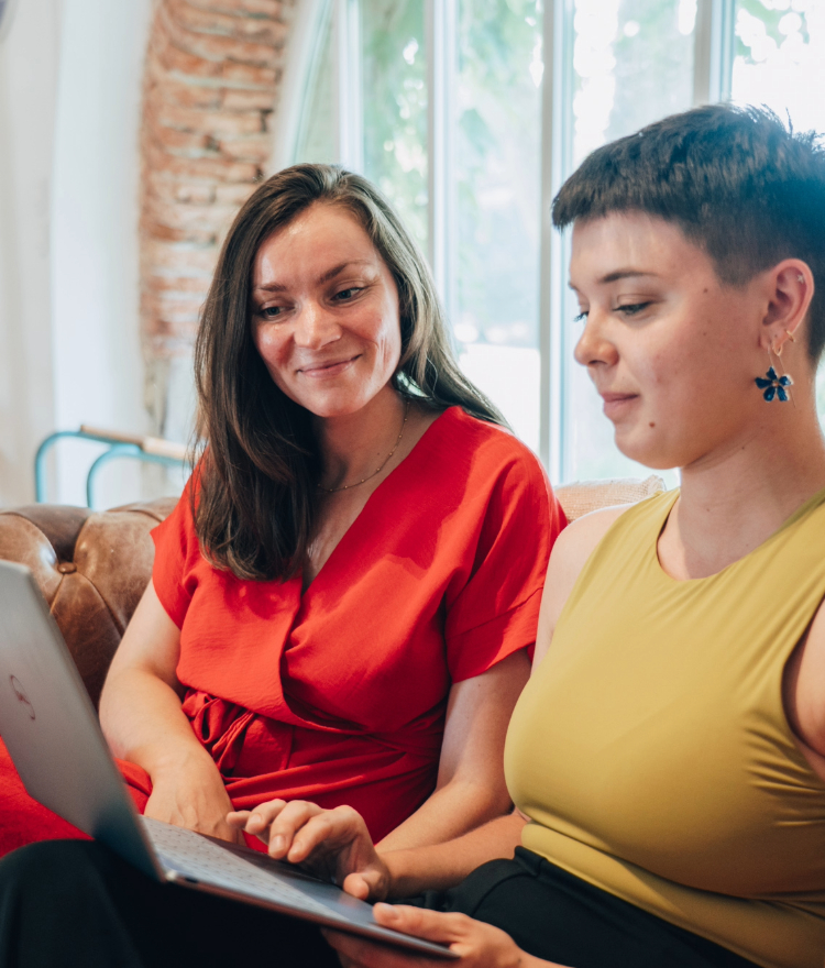 Deux femmes joyeuses et concentrées, assises sur un canapé, tenant un PC et observant attentivement l’écran.
