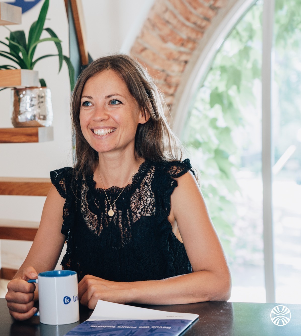 Une femme souriante, assise à une table, avec devant elle un livret et un mug Linklusion.