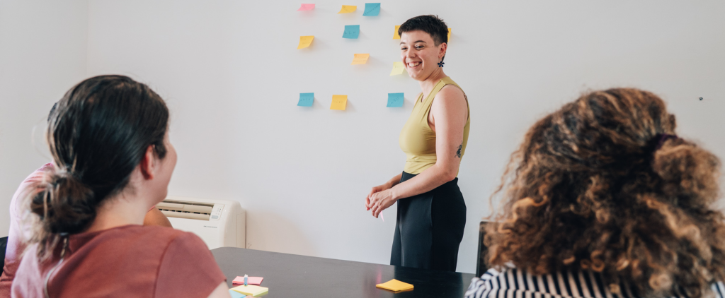 Photo d'une personne debout qui colle des post it sur un mur en face de 3 personnes assises.