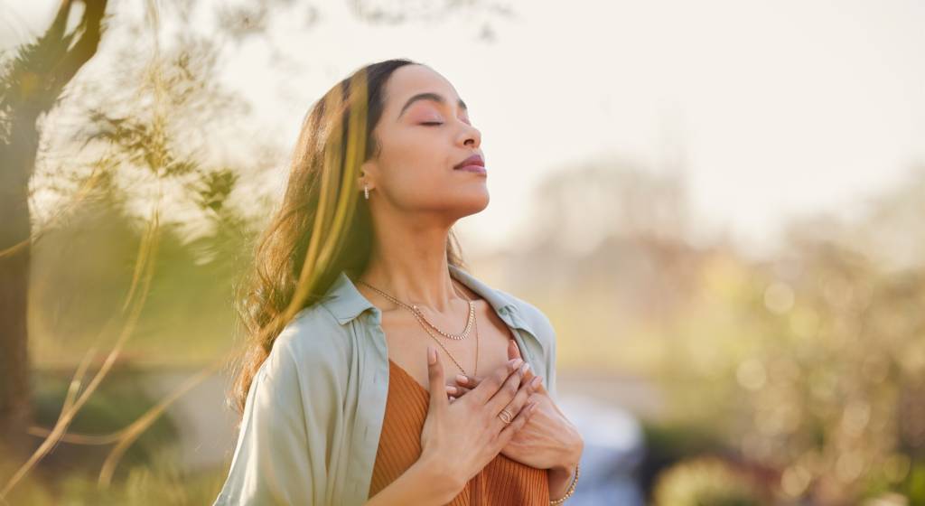 Femme debout, en extérieur qui pose ses mains sur son thorax et qui respire en fermant les yeux.